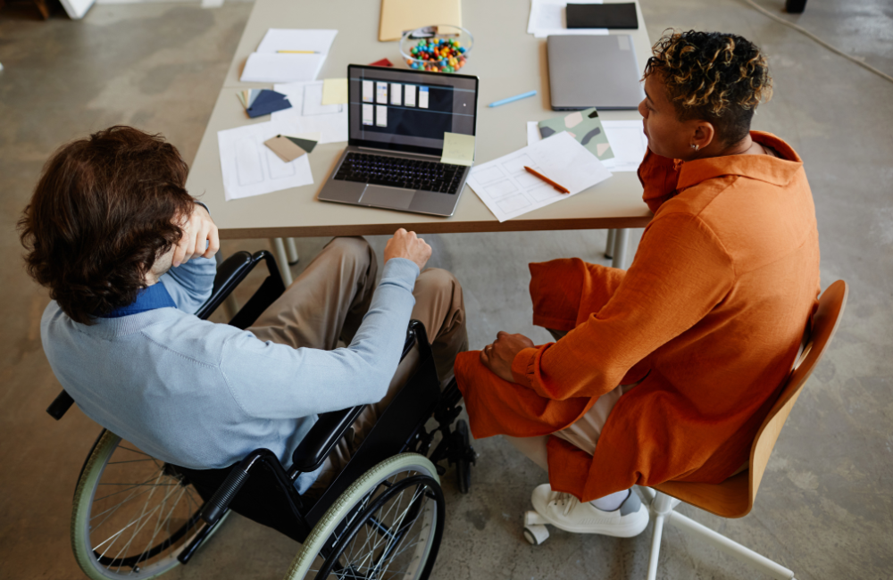 Twee mensen overleggen aan een tafel met laptop; één persoon zit in een rolstoel.