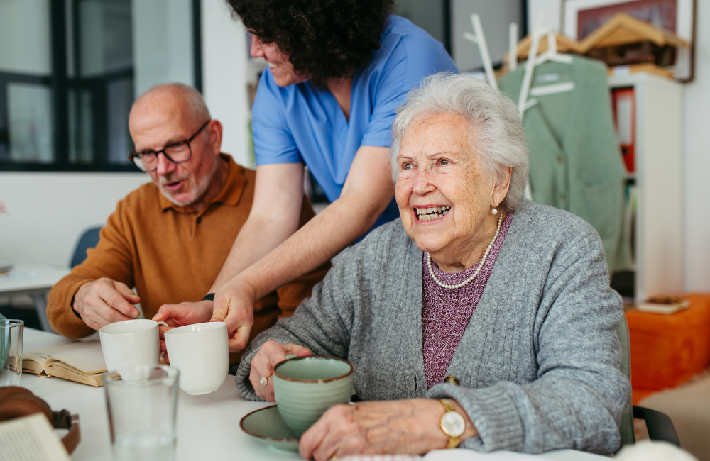 In een woonkamer lacht een vrolijke, oudere vrouw terwijl een verzorger haar en een andere man aan een tafel twee kopjes koffie aanreikt.